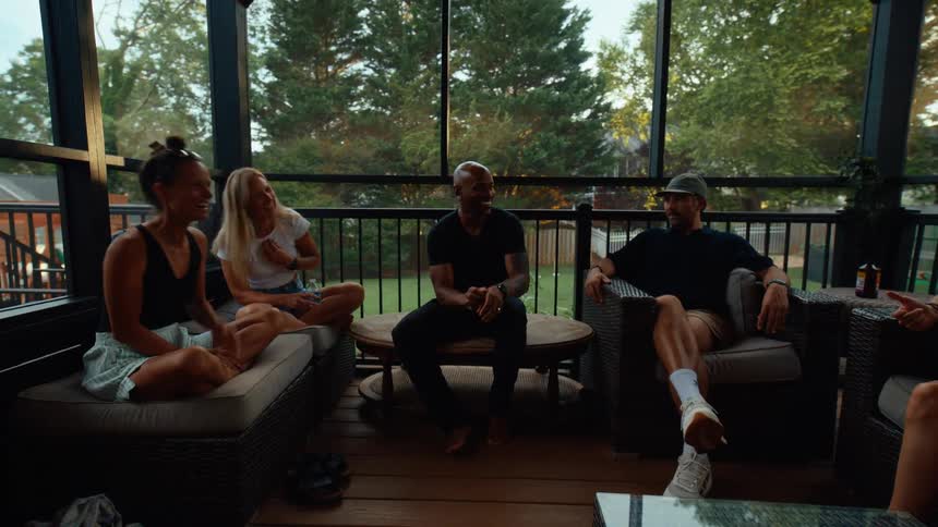 Three adult women and two men sit on chairs and a couch on a porch or patio talking with trees in the background on a sunny evening