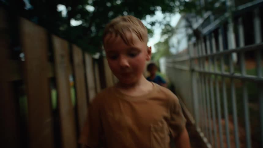 Two young boys run along a narrow path between two buildings bordered by backyard fences in a residential neighborhood at midday