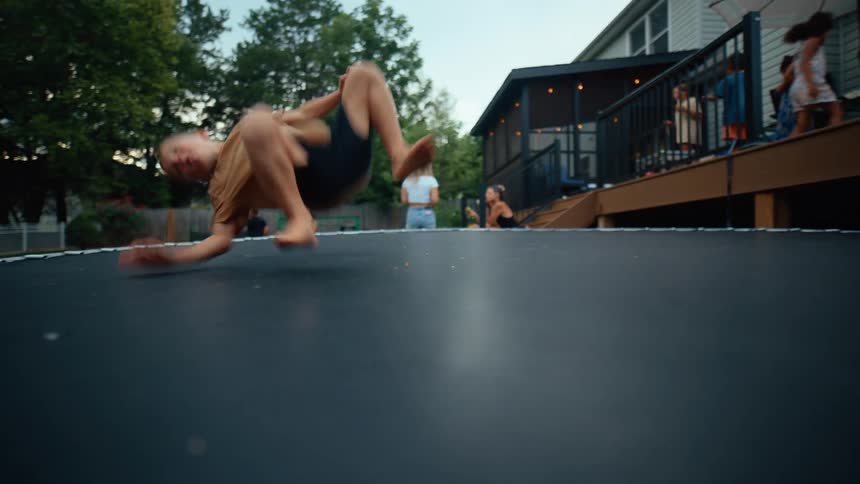 A young boy performs a somersault on a trampoline while people relax on a nearby porch