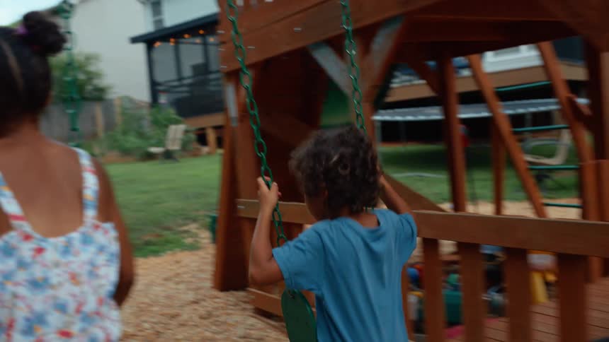 Two children are playing on a swing set in a backyard a woman stands in the background watching them while a man is shown closeup also watching