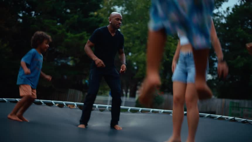 A multiracial family of four jumps and bounces on a large trampoline in their backyard the group is laughing and having fun as they jump up and down