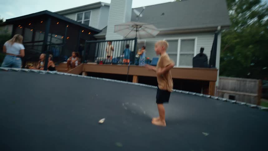 Two barefoot boys jump on a trampoline in a suburban backyard with a family gathered on a large wooden deck in the background under an umbrella