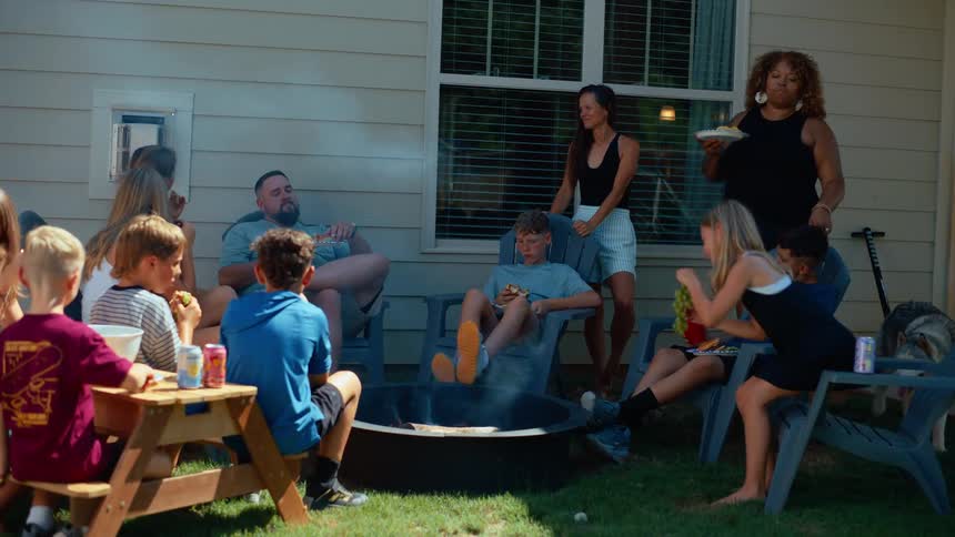 Several kids sit in a backyard with two women and a man having lunch and enjoying time together on a summer day