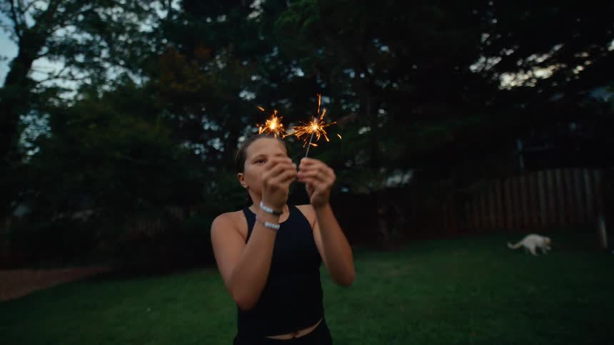 Teenager girl playing with sparklers next to a group of adults and children celebrating in a lawn outdoors during day time