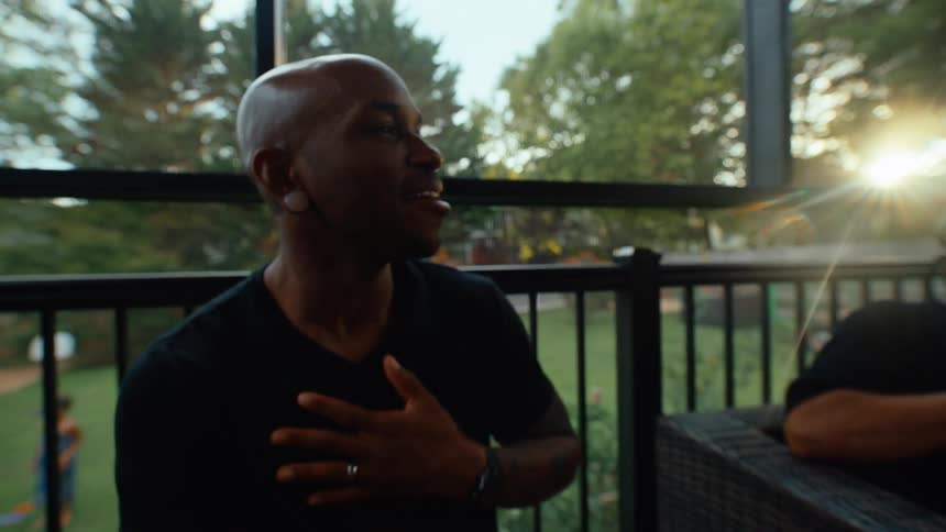 A bald man in a black shirt is sitting on a patio smiling and laughing he is gesturing with his hands and appears to be enjoying a conversation with someone offcamera