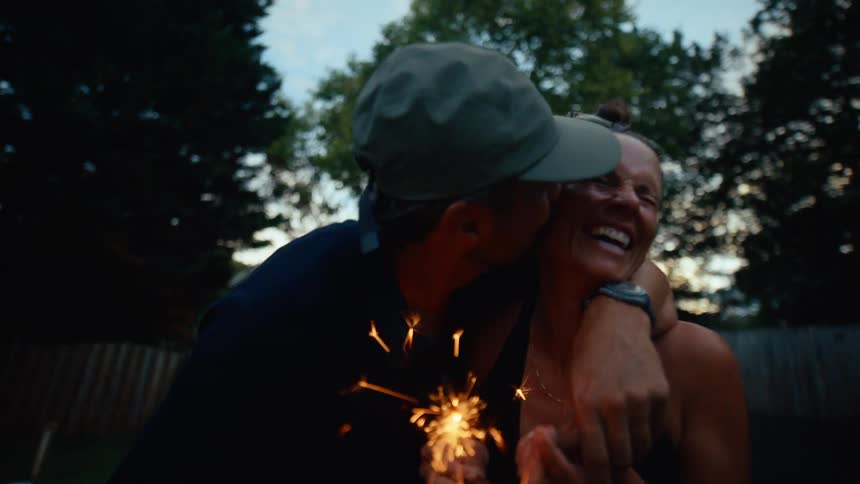 A man wearing a hat stands in a backyard at sunset his hands resting on the shoulders of a young woman both hold discostyle fireworks and laugh excitedly together