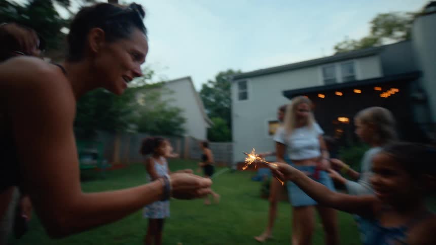 A lively midday backyard gathering where family and friends enjoy time together on a sunny lawn kids laugh and play some holding small discostyle fireworks that sparkle in the daylight while adults chat relax and join in the fun