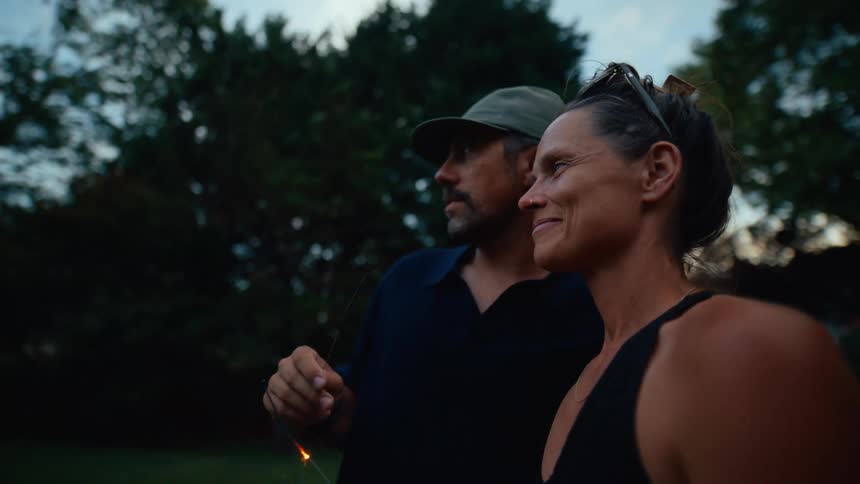 A smiling woman and a man are holding a sparkler in a backyard at dusk with trees and a dark sky in the background