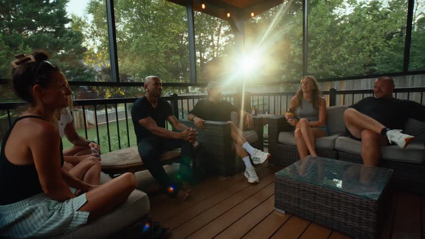 Small group of young adults sitting on sofas and talking on a deck next to a house outdoors during sunset time