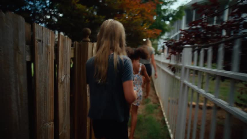 Three female kids walk behind a teen girl on a narrow path at midday with an averageheight wooden fence on the left and a steel fence on the right