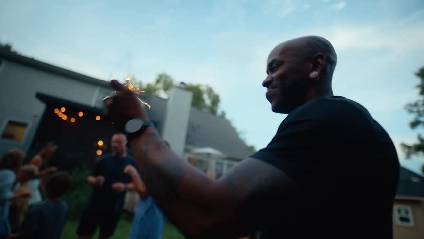 Black man playing with a sparkler surrounded by other adults and children running and laughing in a celebreation at the backyard of a house outdoors by daylight