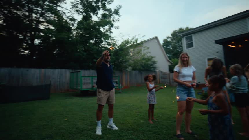 A diverse group of adults and children are celebrating in a backyard at dusk they are standing on the lawn holding sparklers that are lit and sparkling