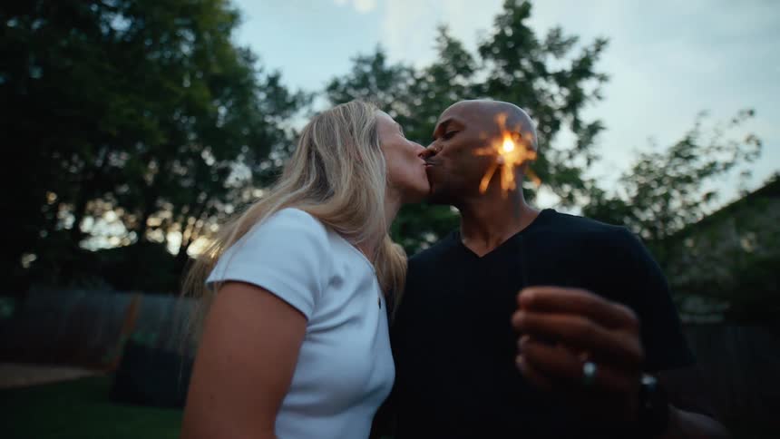 A diverse couple stands close together in a backyard at dusk they are holding lit sparklers and are about to share a kiss