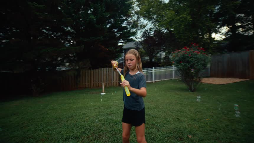 A smiling woman in jean shorts and a boy and a girl are playing with large bubble wands in a sunny backyard with a large house and a fence in the background