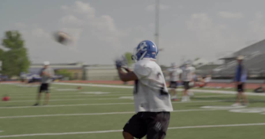 In the video two lines of people are visible on a football field the first line composed of what appear to be football players in blue jerseys is positioned in the foreground another line of what also appears to be players in white jerseys stands in