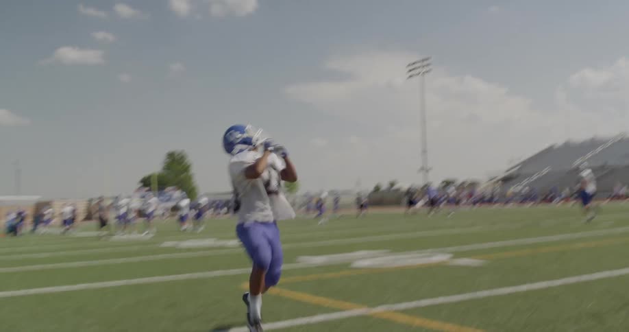 The video shows a football player wearing a white jersey and blue pants running toward the camera with a football in his hands in the background there are two distinct lines of players visible on the field the players in both lines are wearing blue j