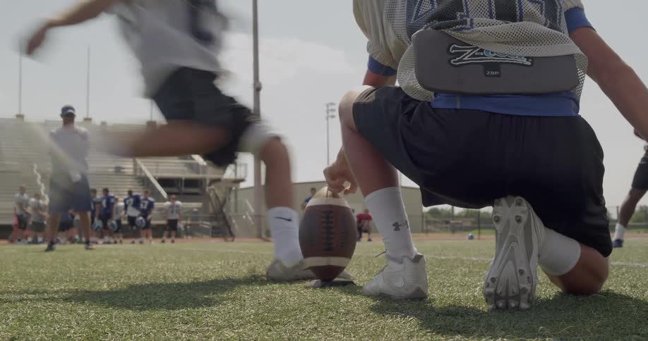 A football player holds a football on a tee while another player kicks it during practice on a sunny field with a coach and team in the background