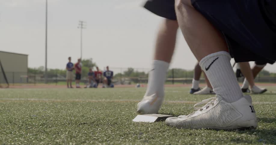 The video shows a lowangle shot of a football training session in the foreground a football player is on one knee holding a football in the background there are two distinct lines of football players one line consists of players in blue jerseys an