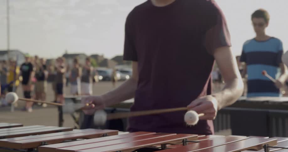 The video shows two lines of people at an outdoor musical practice in the foreground a musician wearing a maroon shirt is playing a marimba with mallets in the background two separate lines of people are standing and facing the foreground the people