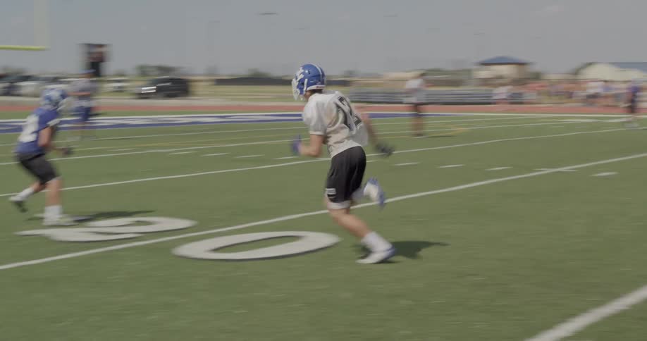The video shows a football practice with two groups of players on a field in the foreground two players are in a drill running from the right side of the frame toward the left in the background two lines of football players are facing each other in a