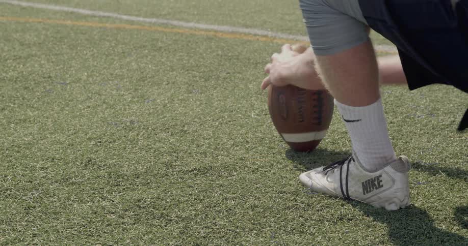 The video shows two distinct lines of football players in uniforms in the foreground a player in a black jersey with a white number is kneeling on the grass with his hand on the ground in the background there is a second line of players also in a cro