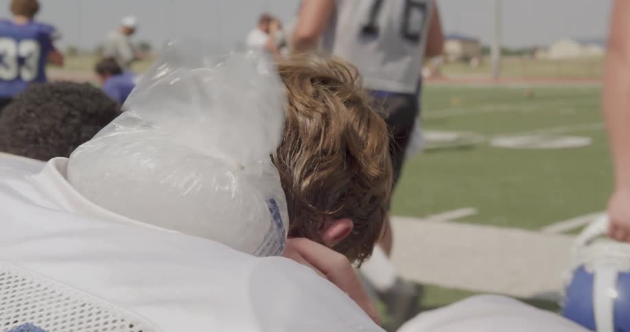American football player sits on a bench near the football field while icing his neck on a sunny day