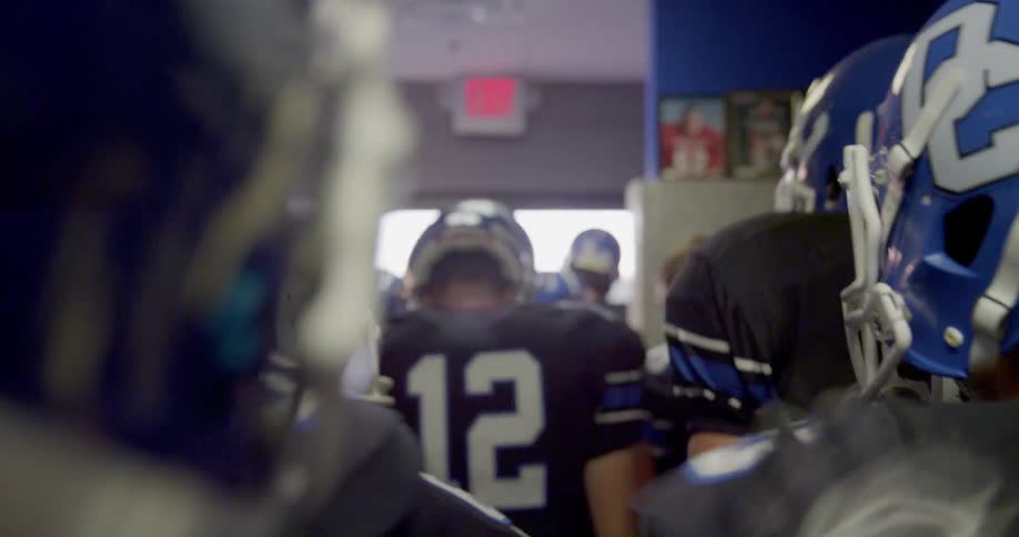 A group of american football players wearing black jerseys and blue helmets stand just inside the stadium entrance at midday looking excited and ready to step onto the field for their match