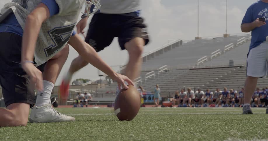 The video shows two lines of people at a football practice in the foreground one person is kneeling and another person is standing with a football on the ground between them in the background two separate lines of people are sitting in the bleachers