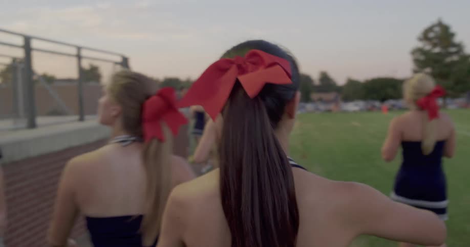 The video shows a group of cheerleaders walking on a field viewed from behind they are wearing navy blue dresses with white trim and have their hair in ponytails with large red bows there are two distinct lines of cheerleaders with one line walking sl