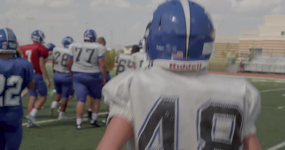 Football players in blue and white jerseys some wearing helmets walk on a field during practice under a sunny sky