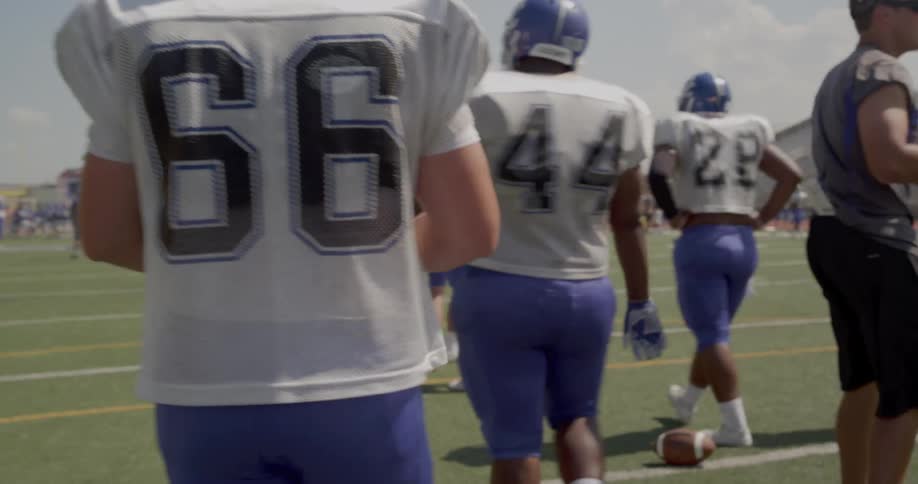 An american football player wearing a white jersey with the number 66 on the back walks among other players on the field under a bright sunny sky