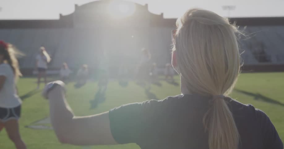 The video shows two lines of people on a field a coach or instructor with blonde hair in a ponytail stands in the foreground with her back to the camera observing the people in the background two distinct lines of cheerleaders both wearing black sho