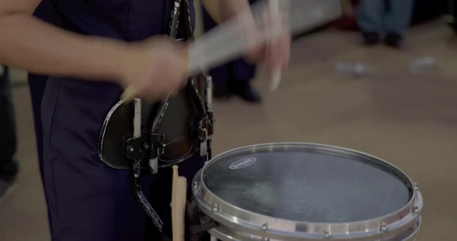 A person in a dark uniform uses drumsticks to play a snare drum with a black head
