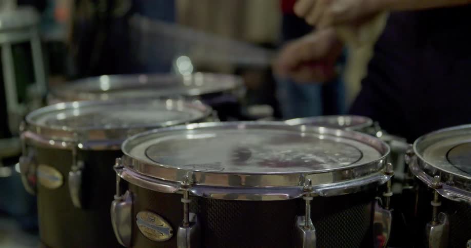 The video shows two lines of musicians with a focus on a drummer in the foreground he is playing a set of drums with drumsticks in the background there is a second line of musicians standing behind the drummer and they appear to be part of a band or