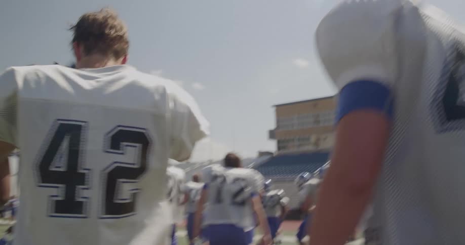 American football players walk off the pitch on a sunny day after a game removing their helmets as they make their way toward the sideline