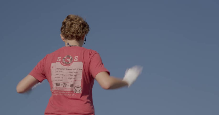 A teenage boy wearing a red tshirt and white gloves stands outdoors in the sun waving his hands left and right