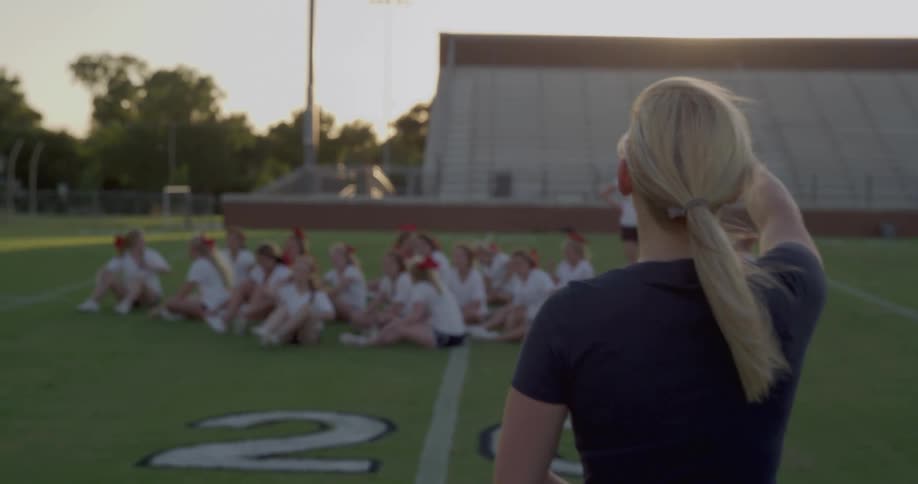 Blonde woman gestures while providing instructions to teenagers sitting on the turf in a football field near sunset