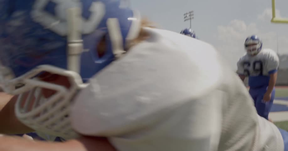 The video shows a football player running a drill on a field he is in a white jersey and blue helmet and is seen in the foreground pushing a blue blocking sled in the background there is a line of other football players in similar uniforms waiting for