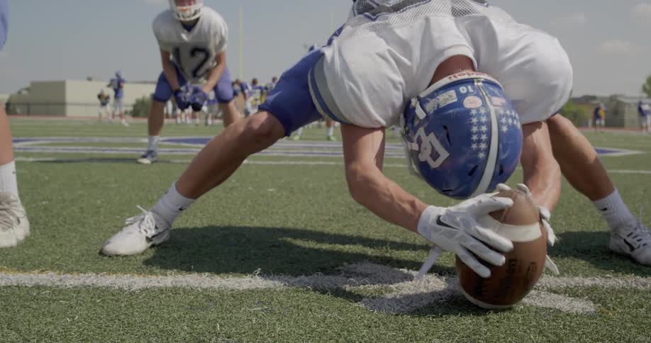 A football player in a blue helmet and white jersey snaps the ball with other players visible in the background on the field