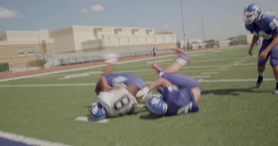 The video shows two distinct lines of football players in an action sequence on a field in the foreground players in a blue uniform are engaged in a drill tackling and running in the background a stationary line of players in white jerseys stands on