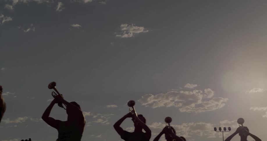 A group of young men stand outdoors on a football field at sunset blowing trumpets as the warm light casts long shadows across the grass
