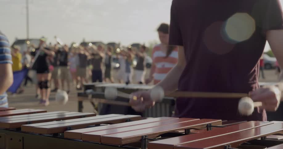 The video shows two distinct lines of people at a musical practice in the foreground a musician is playing a mallet instrument like a marimba with their back to the camera in the background there is another line of musicians standing behind the pers