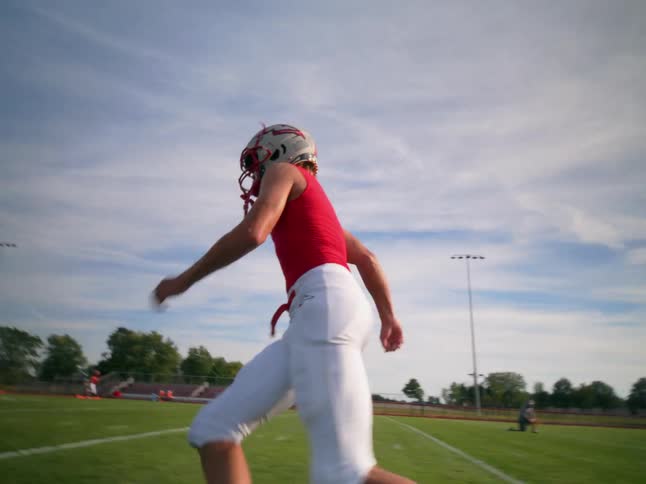 An american football player wearing a tight red shirt and white pants runs around the pitch at midday helmet on under the bright sun