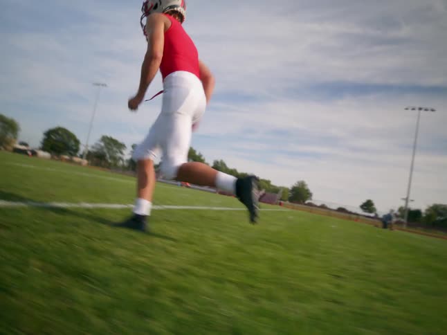 The video shows two lines on a football field a white line is painted on the grassy field in the foreground and a football player in a red jersey and white pants runs along it in the background there is a line of players standing on the field