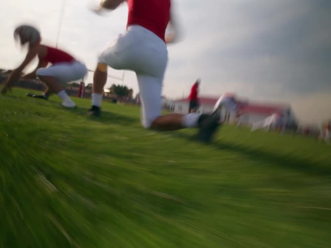 A very low close view shows a male high school football player in a red jersey and white pants stretching on a grassy field during practice
