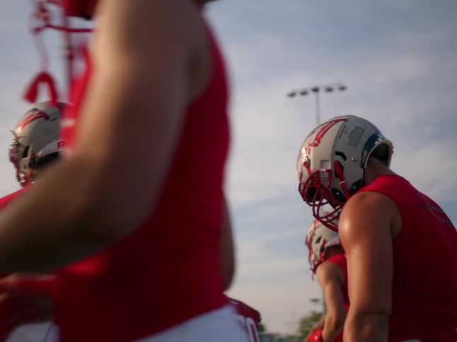 The video shows two distinct lines of football players in uniforms in the foreground one player in a red jersey is out of focus and standing with their back to the camera in the background there is a second line of players also in red jerseys waiting