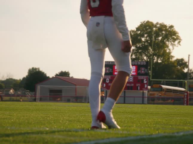 The video shows two distinct lines of people in the background in the foreground a football player in a red jersey with the number 3 is walking on a football field in the background there is a line of school buses further behind the buses