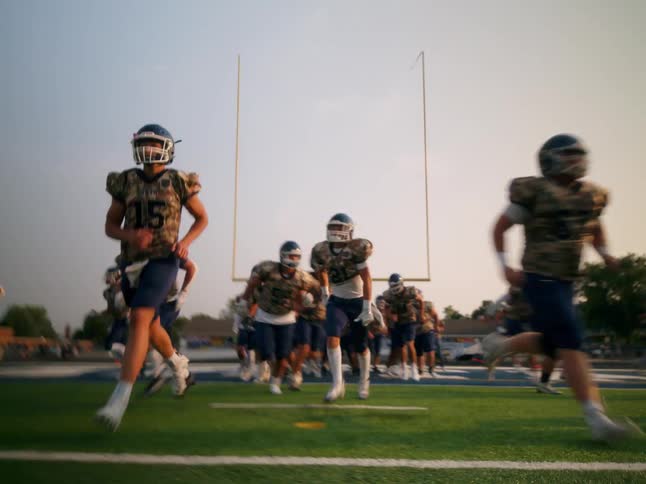 A group of high school football players in full uniform running onto a football field for practice