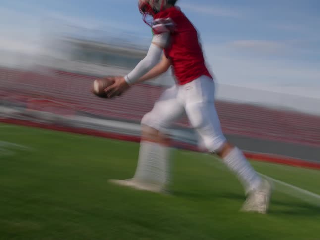 A football player in a red jersey and white pants runs with the ball on a green field