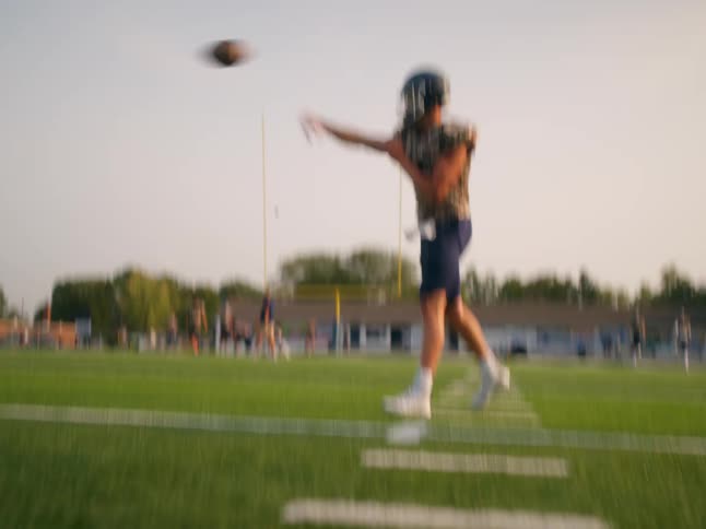 An american football player wearing a helmet and a yellow jersey throws the ball across the field at midday the bright sunlight highlighting the motion as it arcs through the air